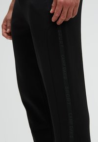 Schwarze Sporthose aus einem weichen Material, mit schlanker Passform, seitlichen Streifen mit dem Text "SPORT CAMP DAVID" und einer glatten Oberflächenstruktur.