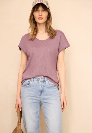 Kobieta ubrana w fioletową bluzkę z krótkim rękawem, jasnoniebieskie dżinsy i wzorzystą czapkę, trzyma brązową torbę na beżowym tle.