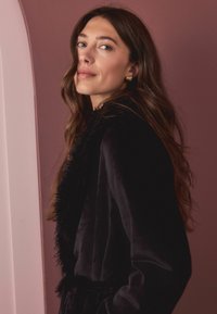 Juodas aksominis chalatas su tekstūruotu kailio apykakle, surištas juosmenyje. Fonas švelniai rožinis, pabrėžiantis chalato spalvų kontrastą.