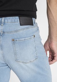 Jeans de denim azul claro con un corte slim, que presentan una etiqueta de piel negra en la cinta de la cintura, un bolsillo trasero de forma redondeada y detalles de costura sutiles.