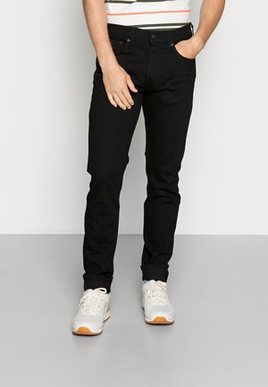 Mężczyzna w czarnych jeansach typu slim-fit i białych trampkach stoi na jednolitym jasnym tle z jedną ręką w kieszeni.