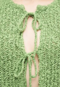 Πλεκτό ένδυμα πράσινης υφής δεμένο με δύο φιόγκους, που αφήνει το δέρμα να φαίνεται μέσω του μπροστινού ανοίγματος.