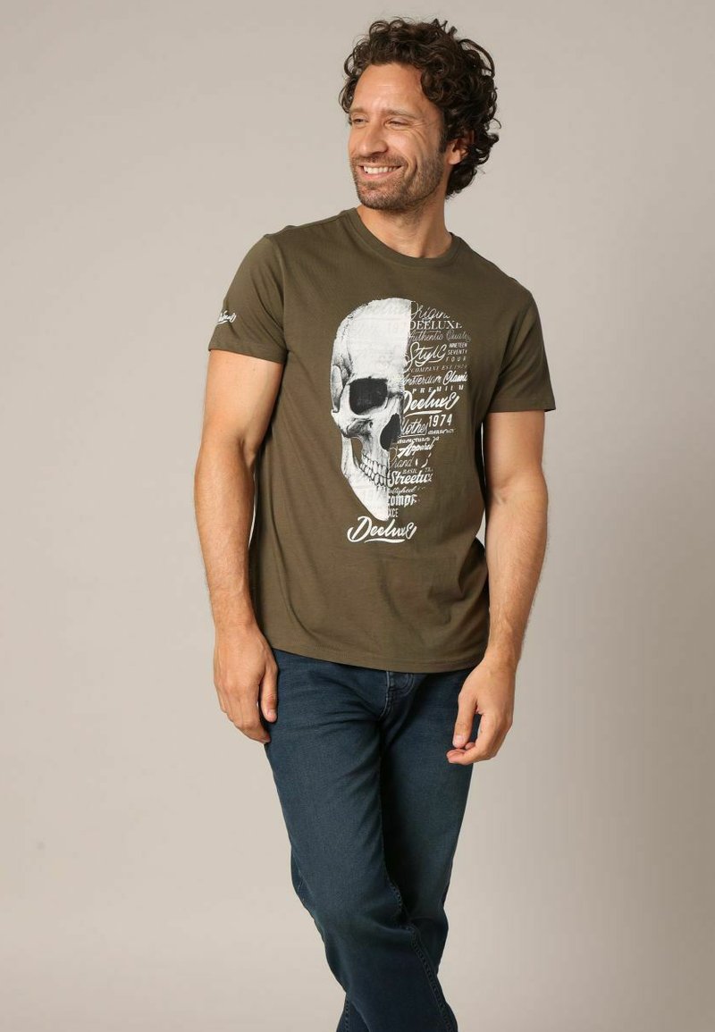 T-shirt vert olive avec un graphisme de crâne blanc et une impression de texte. Manches courtes, encolure ronde et coupe décontractée. Jean en denim associé au t-shirt.