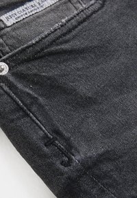 Μαύρο ύφασμα τζιν με ελαφριά γυαλάδα, με ραμμένες λεπτομέρειες, μπροστινή τσέπη και ετικέτα με το λογότυπο κοντά στη ζώνη.