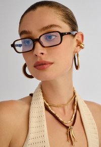 Γυναίκα με μαλλιά χτενισμένα πίσω, φοράει τετράγωνα γυαλιά με φλοίσβο χελώνας, μεγάλα χρυσά σκουλαρίκια κρίκους και στρωμένα κολιέ με χρυσά φυλακτά.