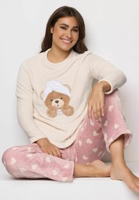 Donna seduta a gambe incrociate che indossa una maglia a maniche lunghe color crema con una grafica di un orso e pantaloni in pile rosa decorati con cuori bianchi.