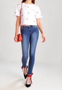Camiseta blanca recortada con bordado floral, combinada con jeans ajustados azules. Tacones negros con acentos de cintas rojas y pequeño bolso cruzado naranja.