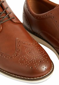 Chaussures en cuir marron avec un design brogue, présentant des détails perforés et une texture lisse, complétées par une semelle blanche contrastante.