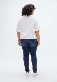 Λευκό κοντομάνικο πουκάμισο συνδυασμένο με σκούρο μπλε στενά τζιν και ανοιχτόχρωμα αθλητικά παπούτσια, από πίσω σε απλό φόντο.