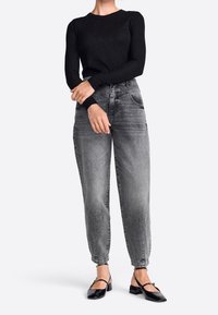 Schwarzes Langarmoberteil mit Rundhalsausschnitt, kombiniert mit lockeren grauen Jeans, die ein leicht verblasstes Finish und abschließende Cuffs an den Knöcheln haben. Schwarze flache Schuhe.