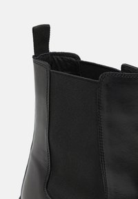 Bottine noire avec une finition en cuir lisse, panneaux latéraux élastiques et une languette pour un enfilage facile. Un tissu texturé contraste avec le cuir.
