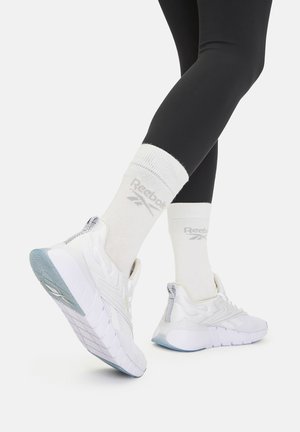 Białe sportowe buty z teksturowaną siateczką i gumową podeszwą, w zestawie z białymi skarpetkami z szarym logo, noszone z czarnymi legginsami.