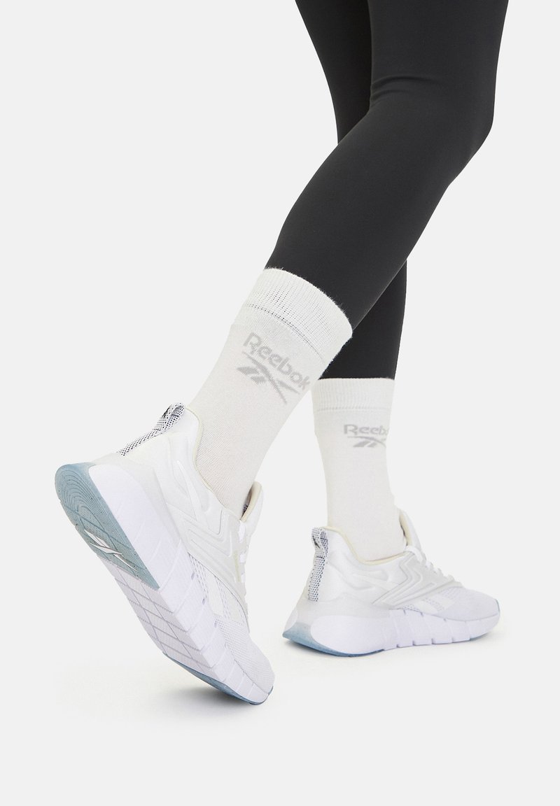 Biele športové topánky s textúrovanou sieťovinou a gumovou podrážkou, v kombinácii s bielymi ponožkami so sivým logom, nosené s čiernymi legínami.