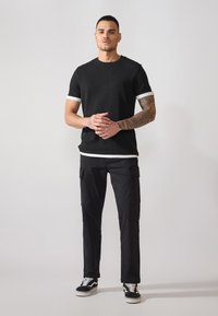 Sort tekstureret kortærmet skjorte med hvide accenter, parret med sorte cargo-bukser og sorte sneakers. Modellen står foran en neutral baggrund.