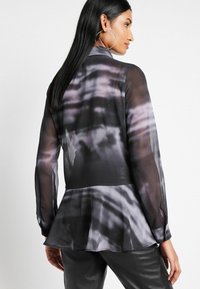 Gennemsigtig lang�ærmet bluse i sort og grå med et tie-dye mønster, der har en peplum-hæm og knaplukning ved håndleddene. Glat tekstur.