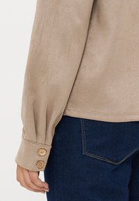 Beige suede-like top featuring a loose fit, buttoned cuff details, and seamless hem. Paired with dark blue denim.