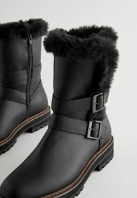 Bottes en fausse cuir noir avec un revers en fourrure, dotées de deux accents de boucles métalliques et d'une semelle texturée. Fermeture éclair sur le côté pour un enfilage facile.