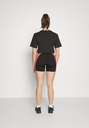 T-shirt noir à manches courtes associé à un short noir avec des panneaux latéraux roses. Le modèle porte des baskets noires avec des chaussettes blanches qui montent jusqu'aux chevilles.