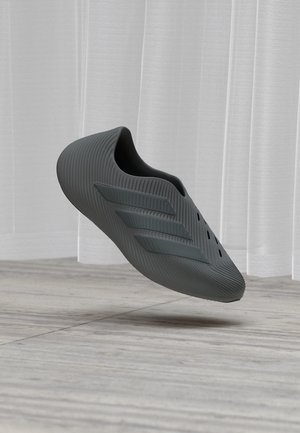 Mørkegrå slip-on-sko med tre hevede striper, ribbet tekstur og ventilasjonshull, svevende over et lyst tregulv.