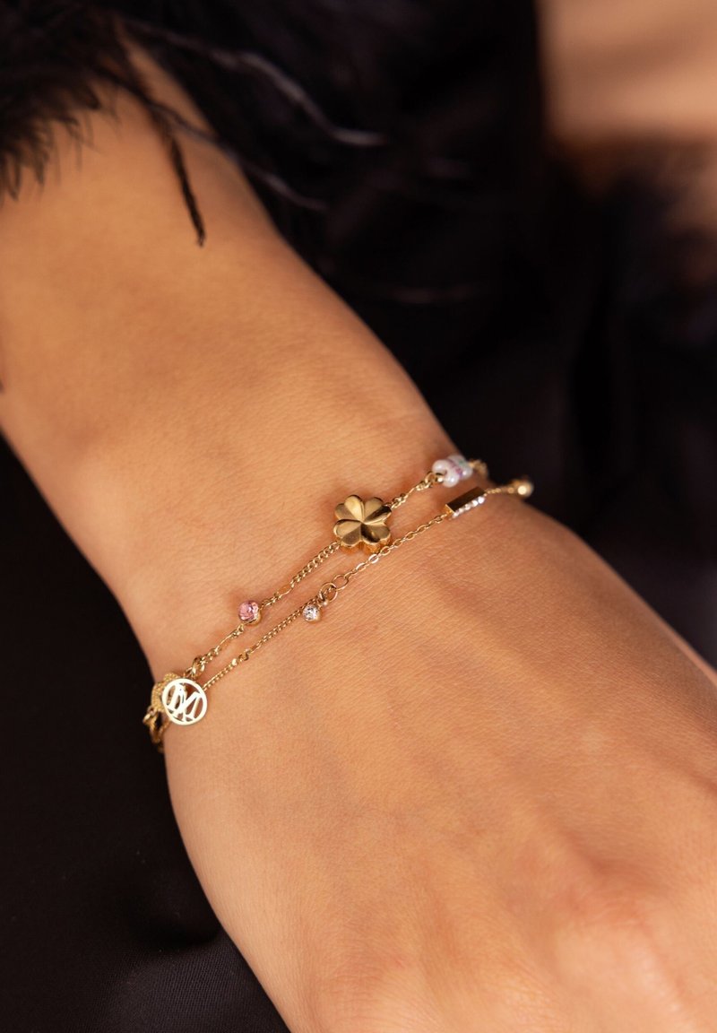 Gouden armband met een bloemenaccent, kleine roze en heldere kralen, gelaagde kettingen en een vredessymbool charm aan een pols met een lichte huidtoon.