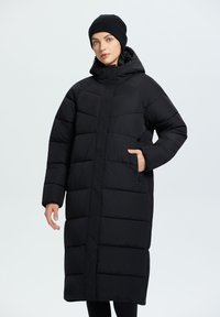 Abrigo acolchado negro con una silueta larga y oversized, secciones acolchadas, bolsillos con cremallera y un cuello alto, llevado con un gorro a juego.