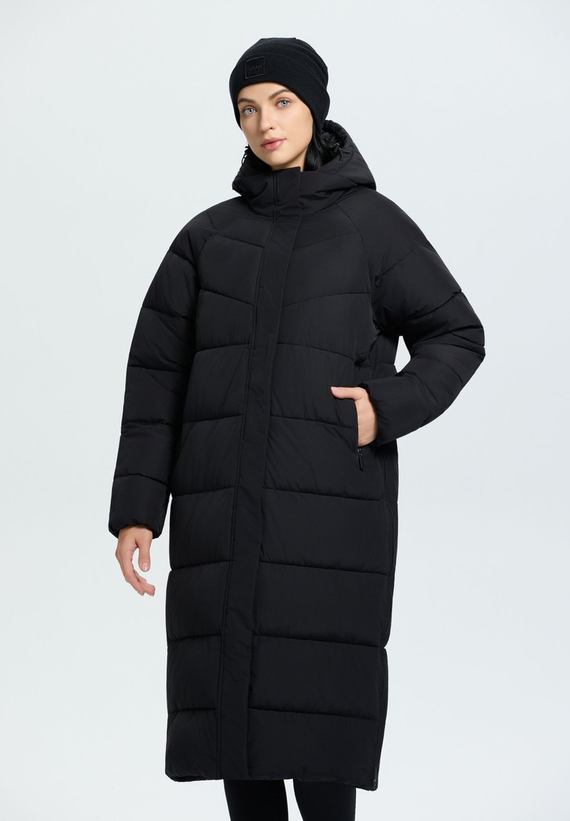 Abrigo acolchado negro con una silueta larga y oversized, secciones acolchadas, bolsillos con cremallera y un cuello alto, llevado con un gorro a juego.