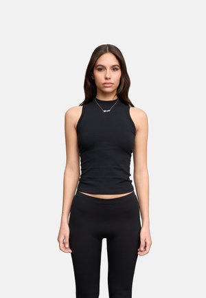 Jonge vrouw met lang bruin haar, gekleed in een mouwloos zwart topje en zwarte legging, staand tegen een effen lichte achtergrond.