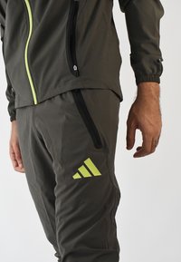 Chaqueta deportiva gris con cremallera y detalles en verde lima, combinada con pantalones a juego que presentan un logo en verde lima y bolsillos laterales con cremallera.