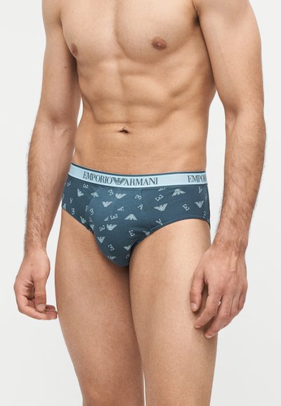 Μυώδης ανδρικός κορμός που φοράει μπλε σλιπ Emporio Armani με λωρίδα μέσης με λογότυπο και σχέδιο με γράμματα και σύμβολα αετού Armani.