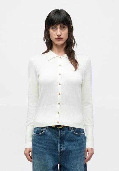 Cardigan blanc avec col, manches longues et boutons dorés sur le devant. Ourlet et poignets côtelés texturés, assortis à un jean bleu.