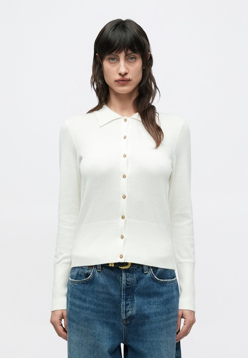 Λευκό ζακέτα με γιακά, μακριά μανίκια και χρυσά κουμπιά μπροστά. Υφή ριμπ στις άκρες και τα μανίκια, συνδυασμένη με μπλε τζιν.