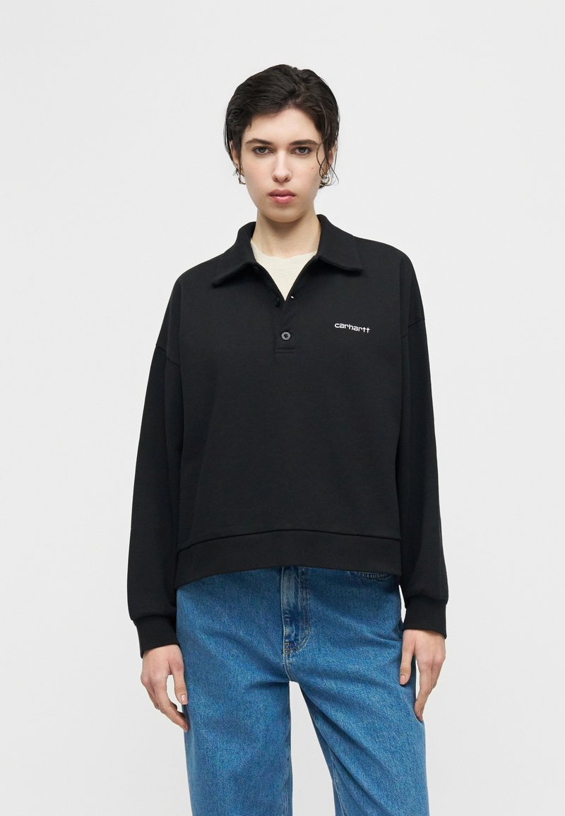 Jeune personne aux cheveux courts et foncés portant un pull Carhartt noir à col et un jean bleu, debout devant un fond clair uni.