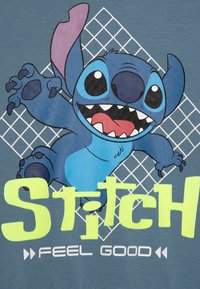 T-shirt graphique bleu représentant un personnage de dessin animé avec de grandes oreilles et des griffes, sur un fond gris avec des lignes de grille blanches. Le texte indique "STITCH FEEL GOOD."