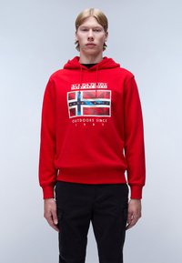Sudadera capucha roja con un estampado gráfico que presenta un diseño de bandera. Puños y dobladillo acanalados. Modelo combinado con pantalones negros. Fondo simple.