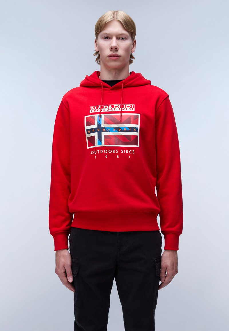 Sudadera capucha roja con un estampado gráfico que presenta un diseño de bandera. Puños y dobladillo acanalados. Modelo combinado con pantalones negros. Fondo simple.