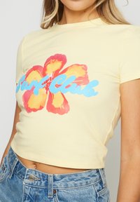 Top corto amarillo hecho de algodón suave, con un gráfico de una gran flor hibisco roja y amarilla y el texto azul "Surf Club" en el centro.