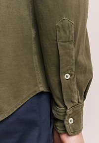 Camicia verde oliva con tessuto strutturato, chiusure con bottoni sui polsini e orlo curvo, con cuciture visibili sui bordi.