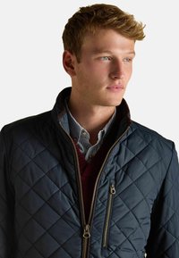 Námornícky prešívaný kabát s vysokým golierom, dvoma zipsovými vreckami a hnedými akcentmi. Nosený cez červený sveter s výstrihom do V a pruhovanú košeľu.