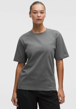 Mujer con el pelo recogido, vistiendo una camiseta gris lisa de manga corta y pantalones negros, de pie frente a un fondo claro y sencillo.