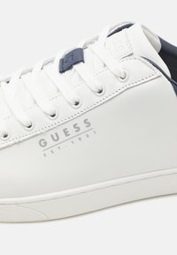 Sneaker in pelle bianca con dettagli blu navy, caratterizzata da punta rotonda, chiusura con lacci e logo embosso "GUESS EST. 1981" sul lato.