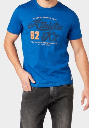 T-shirt en coton bleu avec un imprimé graphique "Athletic Kit 62" en orange et noir, col rond et manches courtes, associé à un jean foncé.