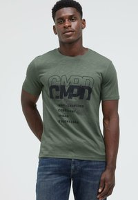 Man wearing olive green CMPD graphic t-shirt and blue jeans, standing with one hand behind back and neutral expression.