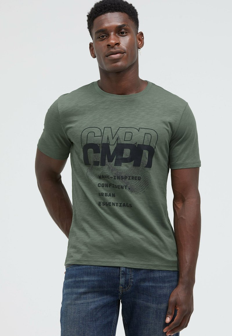 Man wearing olive green CMPD graphic t-shirt and blue jeans, standing with one hand behind back and neutral expression.