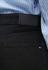 Calças de ganga pretas com um rótulo de couro com a inscrição "TOMMY HILFIGER", apresentando um pequeno emblema de bandeira tri-color na bolsa de trás.