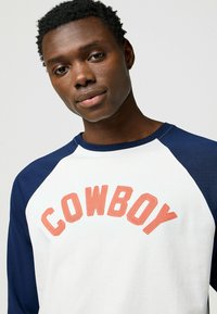 Weißes und marineblaues Crewneck-Shirt mit Raglanärmeln. Mit der Aufschrift "COWBOY" in korallenfarbener Schrift über der Brust. Aus Baumwollmaterial.