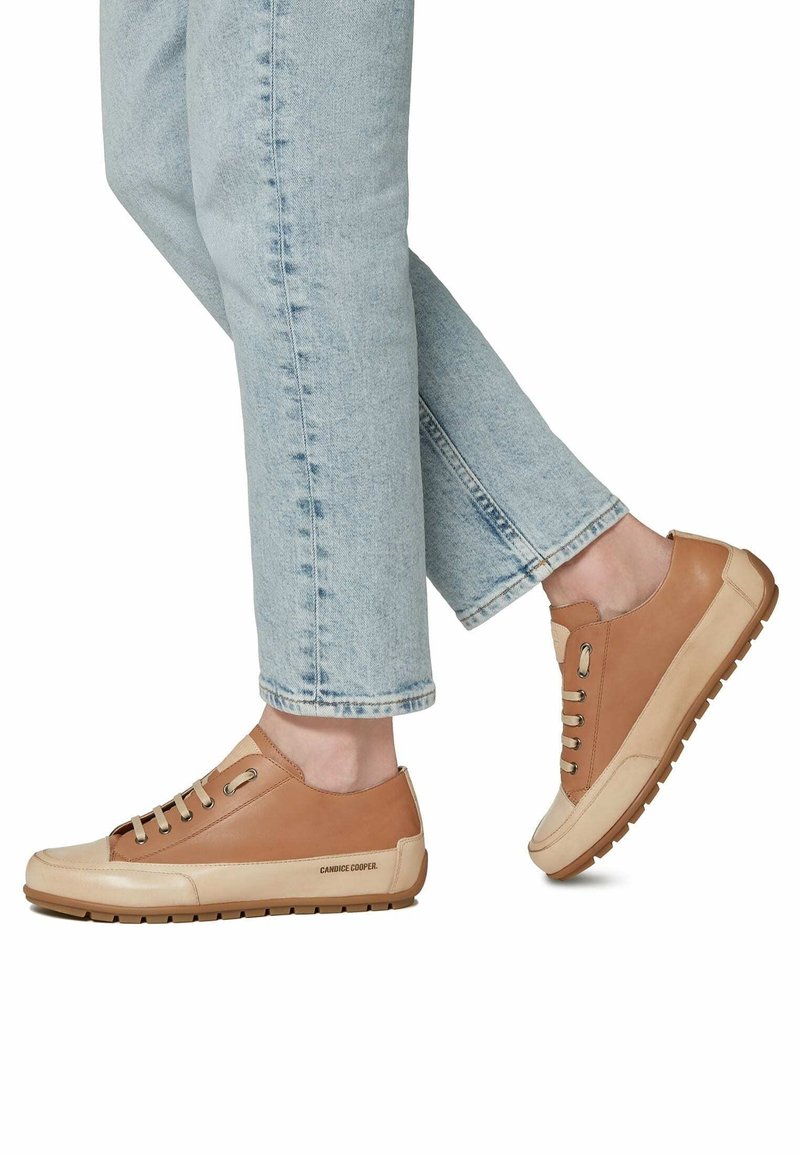 Personne portant un jean bleu clair et des baskets basses en cuir marron avec des semelles et lacets beige, debout sur fond blanc.