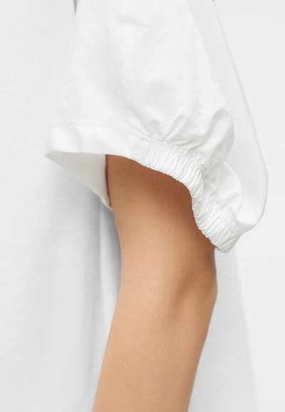 Λευκό κοντό μανίκι με φουσκωτό σχέδιο και ελαστική μανσέτα σε χέρι ατόμου, πάνω σε φωτεινό φόντο.