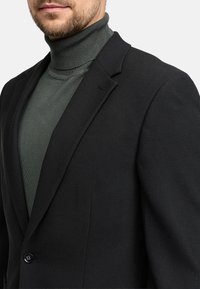Schwarzer strukturierter Blazer mit Revers und einem Knopfverschluss, über einem figurbetonten, dunkelgrünen Rollkragenpullover getragen.