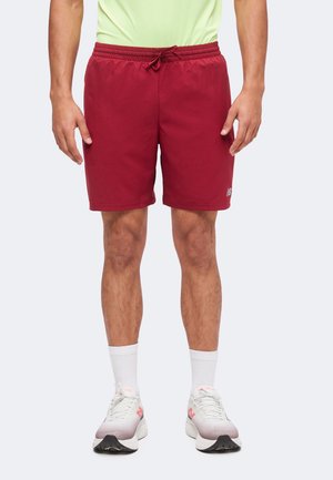 Männlich trägt rote Sportshorts, weiße Socken und weiße Turnschuhe und steht vor einem einfarbigen Hintergrund, Vorderansicht von der Taille abwärts.