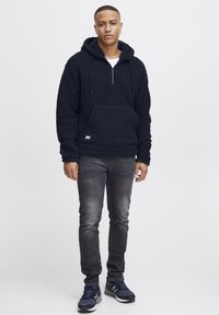 Svart fleece hettegenser med halv glidelås, frontlomme og justerbar hette. Kombinert med mørke jeans og blå joggesko.